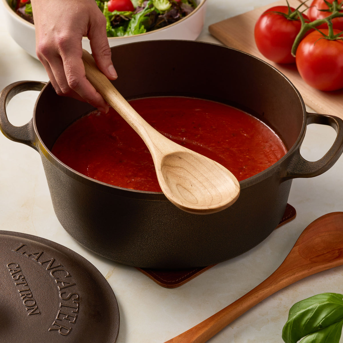 A cast iron Dutch oven pot filled with red tomato sauce, with a hand holding a wooden spoon inside the pot. The lid is shown in the foreground, and fresh tomatoes, basil, and a salad are visible around it on a kitchen counter.